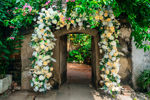 An enchanting floral archway with white and yellow flowers creates a romantic entrance to a lush garden area near Hanoi, Vietnam.