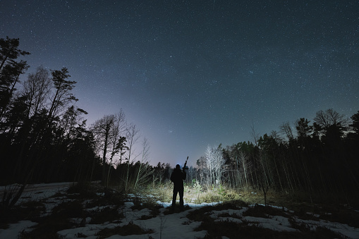 Silhouette of a hunter with a rifle with a silencer and a headlamp in the forest in winter with a starry sky. High quality photo