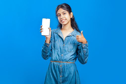 Image of an Indian girl showing cell phone with empty display and ok hand gesture, isolated on blue background