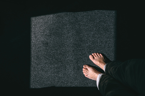 Barefoot male standing on grey carpet, lit by the sunlight from the room window, pov shot top view