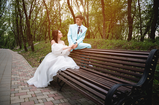 European bride and groom are sitting on a park bench. Newlyweds admiring each other while walking through a green summer park.