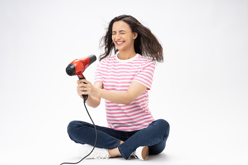 Shot of a beautiful young woman blowdrying her hair while sitting again a white background