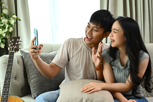 Happy young couple resting on couch taking selfies with smartphone together.