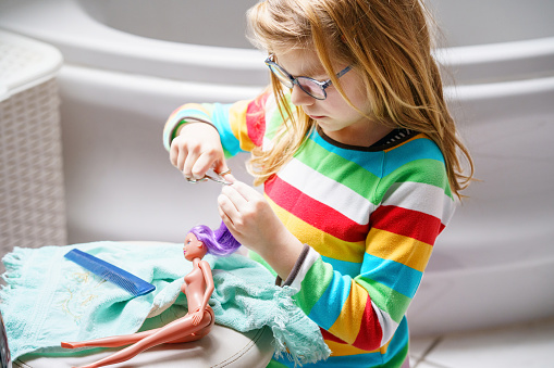 Cute little girl playing with doll at home. Child with eyeglasses making haircut to her toy doll. Indoor creative activities for children.