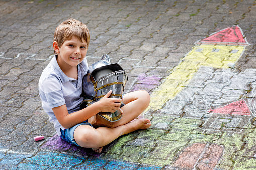 Little active kid boy drawing knight castle and fortress with colorful chalks on asphalt. Happy child with big helmet having fun with playing knight game and painting.