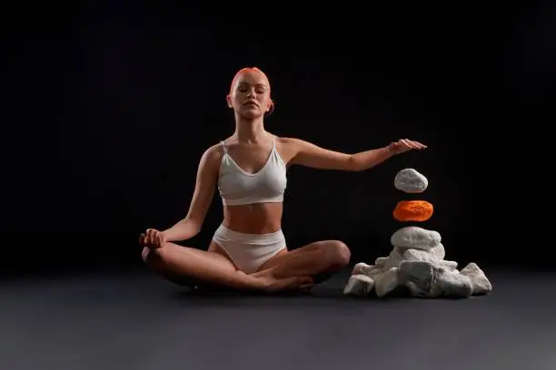 Full body of barefoot female in sportswear sitting in lotus pose and making the rock levitating against black background
