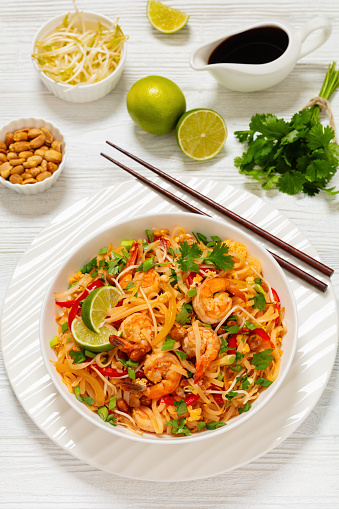 pad thai, phad thai, stir-fried rice noodles with shrimps, peanuts, scrambled eggs, bean sprouts, spring onion, red pepper, sauce, lime in white bowl with chopsticks on wooden table, vertical view