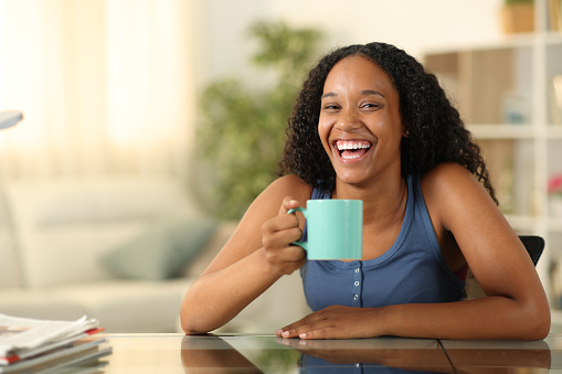 Happy black woman drinking and laughing looking at you at home