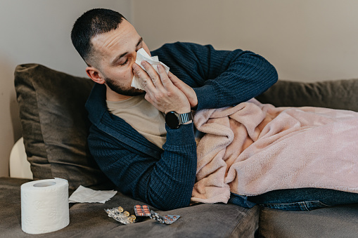 At home, a man faces the discomfort of a cold head-on, blowing his nose with resilience, each action a testament to his determination to overcome the ailment and restore his well-being
