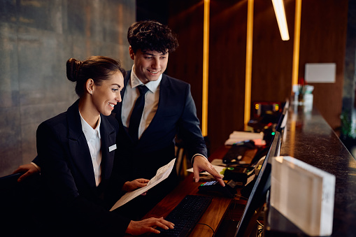 Happy hotel managers through reservation list on desktop PC at reception desk.