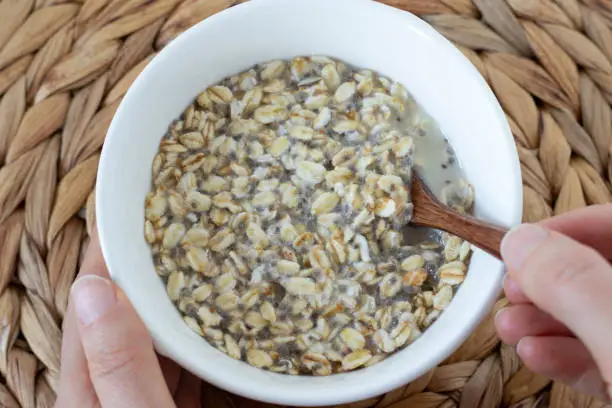 Female hands holding and eating from oatmeal and chia seeds breakfast bowl placed on a bamboo serving tray. Top table view. Close-up. Healthy dieting, sustainability, vegan food concept.