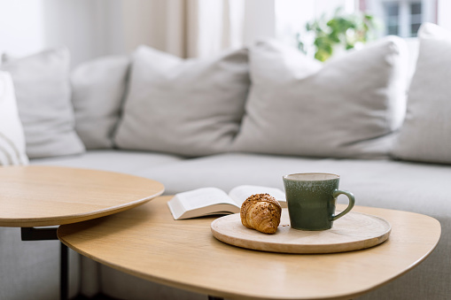 Selective focus on wooden breakfast tray with cup of tea and fresh french croissant on coffee table near comfort sofa with cushions in living room. Rest and relax at cozy apartment