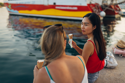 A look over the shoulder at two best friends sitting on the dock, eating ice cream in a cone and casually chatting