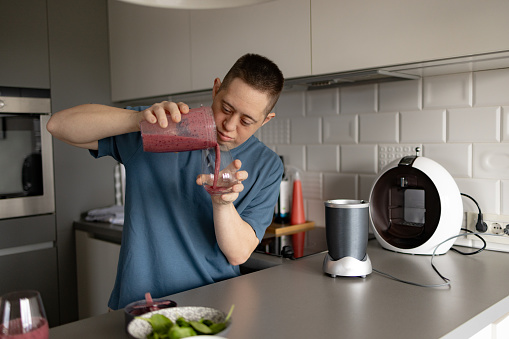Young man with Down syndrome pouring healthy smoothie into a glass in the kitchen.