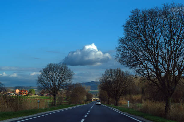 A scenic view of a straight rural road surrounded by green fields and extending towards the horizon under a clear blue sky adorned with fluffy white clouds. Photographed in a Danish countryside setting.