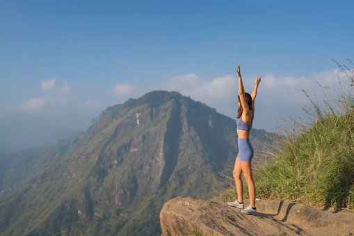 Young woman on top of mountain looks out and raises arms