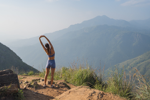 Young woman stretches on top of mountain and looks across valley