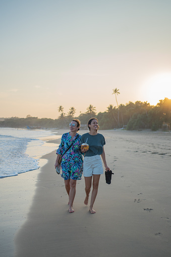Mother and daughter walk down beach together at sunset