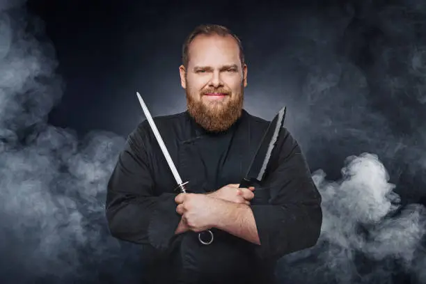 Friendly chef cook holds a kitchenknife and knife sharpener over dark background with lots of smoke. Ready to cook