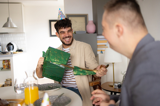 Cheerful man having fun while talking to his friend and opening his gift during Birthday celebration at home.