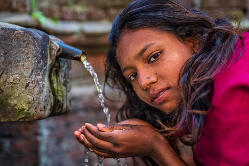 Young Nepali girl drinking from city fountain in Bhaktapur Nepal. Bhaktapur is an ancient town in the Kathmandu Valley and is listed as a World Heritage Site by UNESCO for its rich culture, temples, and wood, metal and stone artwork.