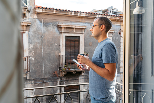 Young man using his smart phone while drinking coffee on the balcony in the morning in Catania In Sicily.