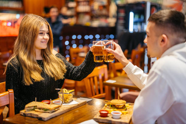 young couple cheering with beer while eating burgers and sandwiches in a pub - toast portion club sandwich cafe photos et images de collection