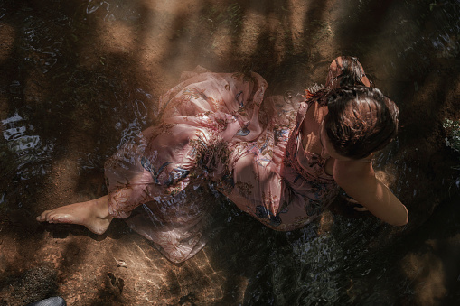 Overhead shot of barefoot young woman in long pink dress enjoying a refreshing bath in the forest stream.