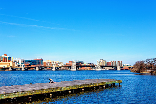 Boston, Massachusetts, USA - March 14, 2024: A woman sitting on a boat dock of the Charles River Esplanade with the Longfellow Bridge and Cambridge shoreline in the background. The Longfellow Bridge is a steel rib arch bridge spanning the Charles River to connect Boston's Beacon Hill neighborhood with the Kendall Square area of Cambridge, Massachusetts. The bridge is also known to locals as the Salt-and-Pepper Bridge due to the shape of its central towers.