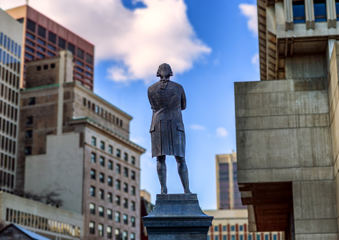 Boston, Massachusetts, USA - March 16, 2024: The Samuel Adams statue on the Faneuil Hall Plaza viewed from the rear, looking towards buildings around the Government Center area. The bronze statue on a granite pedestal was created in 1880 by American sculptor Anne Whitney (1821-1915). Samuel Adams (1722-1803) was an American Patriot who helped organize the American Revolution, signed the Declaration of Independence, and became Governor of Massachusetts. Selective focus..