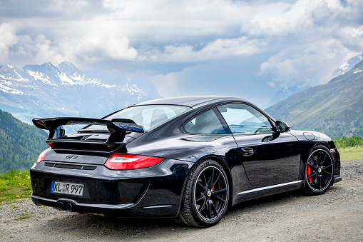 Porsche 911 GT3 sports car (generation 997) in black with red brake calipers against a mountain landscape in the Alps. The Porsche 997 generation was produced from 2004 to 2012, and the GT3 variant is a high-performance lightweight version known for its track-focused capabilities. It features enhancements to suspension, brakes, aerodynamics, and other performance-oriented components. The 997 GT3 is equipped with a naturally aspirated flat-six engine.
