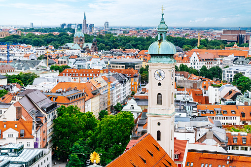 An aerial view of Munich cityscape adorned with historical architecture. The dense arrangement of buildings features red rooftops, each distinct in shape and texture. Streets intersect between the structures, leading the eye through the urban labyrinth. The cloudy sky casts a soft light.