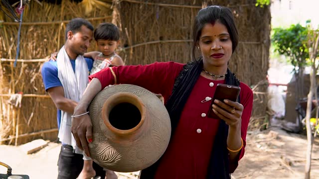 Traditional rural indian woman carrying mud pot while using smartphone in village.