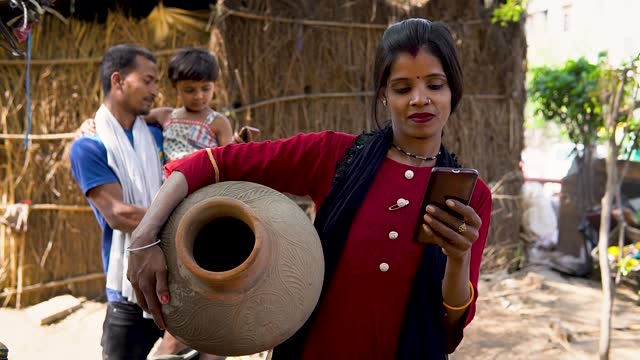 Traditional rural indian woman carrying mud pot while using smartphone in village.