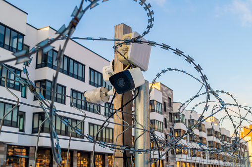 A fence with barbed wire and a camera on top of it. The camera is pointed at a building
