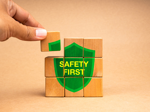 Safety first concept. Hand holding a last piece, put on the wooden cube block puzzle stack with text SAFETY FIRST on green shield icon isolated on brown background. Secure rules symbol at workplace.