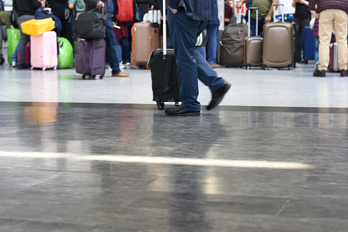 The polished airport floor reflects a traveler walking with a black, upright suitcase, with other passengers and their colorful luggage nearby.