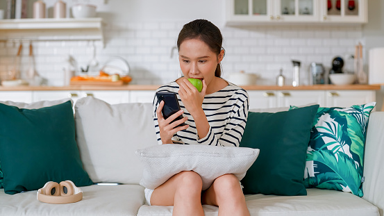 smiling casual asian adult woman relax sit hand hold fresh fruit apple while using smartphone surfting social media online social trend on sofa in living room at home happiness carefree woman sunday lifestyle