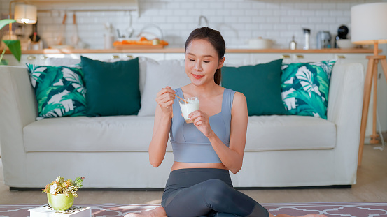 Attractive fitness sport asian woman relax after workout hand hold glass of yogert protein milk shake vitamins after online training exercise course in living room at home good vibe lifestyle