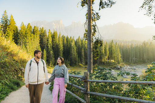 Front view of female with long hair and a male hiking with idyllic view of mountain lake surrounded by pine forest and scenic peaks during shiny sunset in Dolomites Alps, Italy