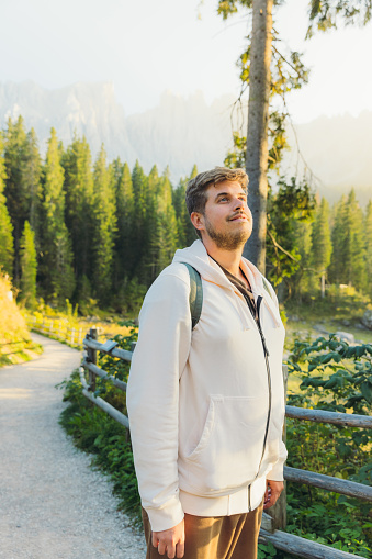Portrait of smiling male with backpack admiring idyllic view of mountain lake surrounded by pine forest and scenic peaks during shiny sunset in Dolomites Alps, Italy