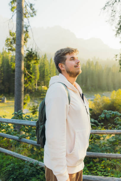 Portrait of smiling male with backpack admiring idyllic view of mountain lake surrounded by pine forest and scenic peaks during shiny sunset in Dolomites Alps, Italy