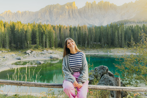 Front view of happy female with long hair relaxing with idyllic view of turquoise mountain lake surrounded by pine forest and scenic peaks during shiny sunset in Dolomites Alps, Ital