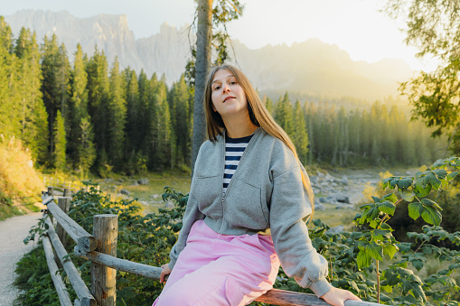 Portrait of female with long hair relaxing with idyllic view of mountain lake surrounded by pine forest and scenic peaks during shiny sunset in Dolomites Alps, Italy