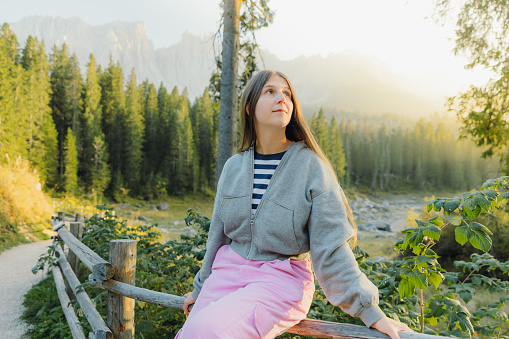 Portrait of female with long hair relaxing with idyllic view of mountain lake surrounded by pine forest and scenic peaks during shiny sunset in Dolomites Alps, Italy