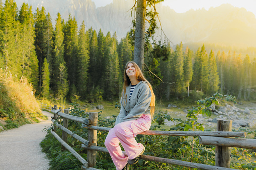 Happy female with long hair relaxing with idyllic view of mountain lake surrounded by pine forest and scenic peaks during shiny sunset in Dolomites Alps, Italy