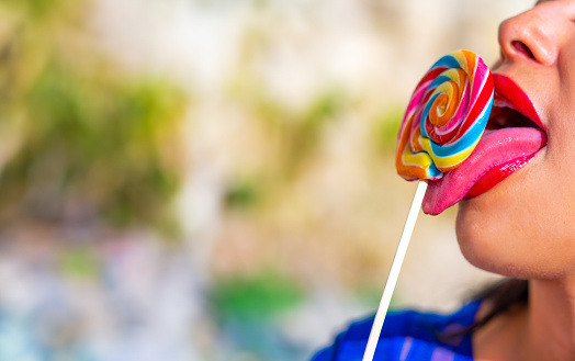 Close up view of bright red full lips and a beautiful long pink tongue sticking out with open mouth tasting a colorful spiral lollipop with copy space.