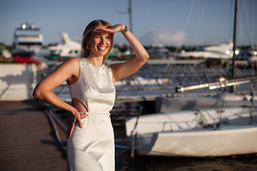 Woman Fashion model strikes a pose on a pier near water with yachts, adorned in luxurious attire long white dress.