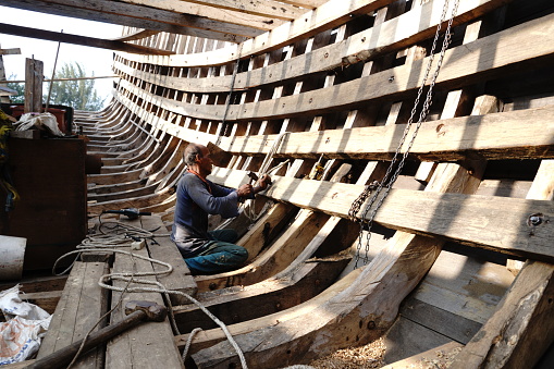 Pak Asbahari, a 54-year-old boat craftsman, is seen here meticulously crafting a wooden fishing boat along the banks of the Krueng Aceh River, Banda Aceh, Indonesia, on August 3, 2023. The construction of wooden fishing boats is a crucial activity in the fishing industry. Workers involved in boat making must possess specialized skills in wood processing and precise assembly to ensure the vessels function effectively and safely at sea. Working alone from dawn till dusk, Pak Asbahari relies solely on his expertise honed over years. Spending seven months crafting this vessel since its inception, he anticipates its completion by the end of 2024.