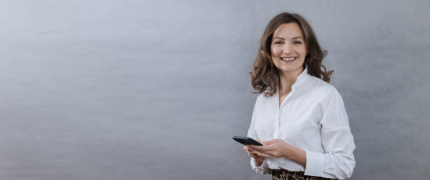 istock Young woman holding a smartphone in her hands, on a gray background, looking at the camera, banner 2094459687
